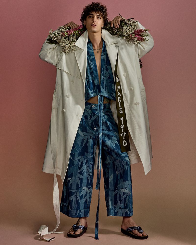 A young adult male fashion model with curly hair poses in a studio wearing a patterned outfit, flowers on his shoulders, and a trench coat.