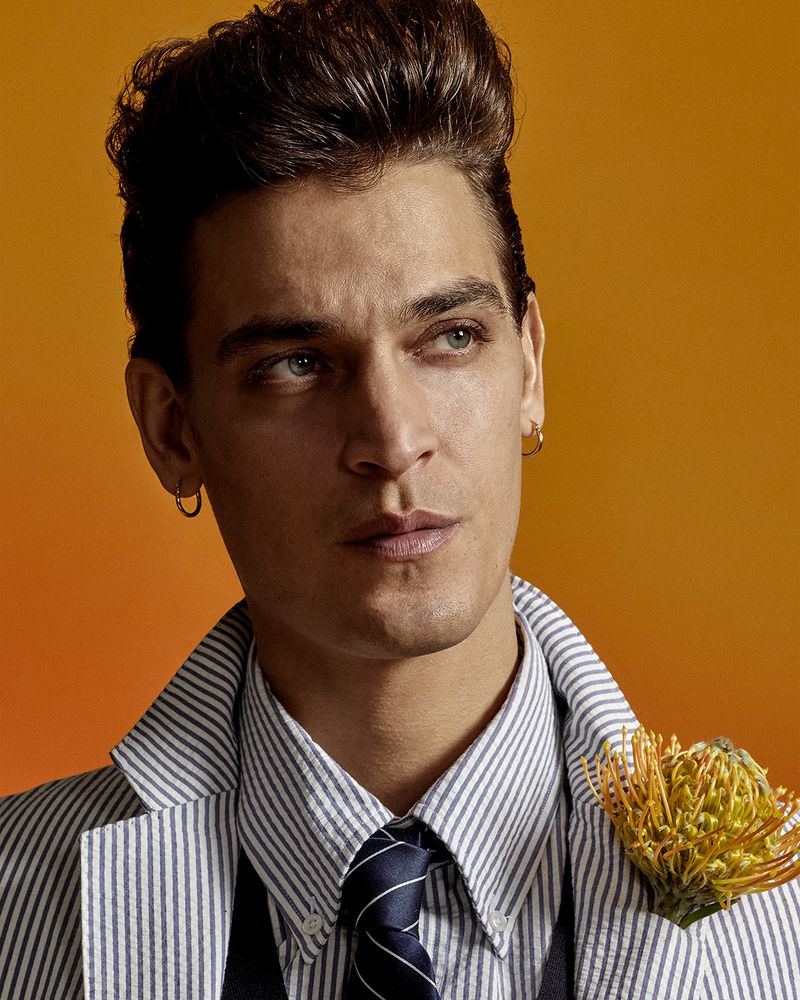 Adult male in a pinstripe jacket and navy tie poses in front of an orange gradient background with a flower in his lapel.