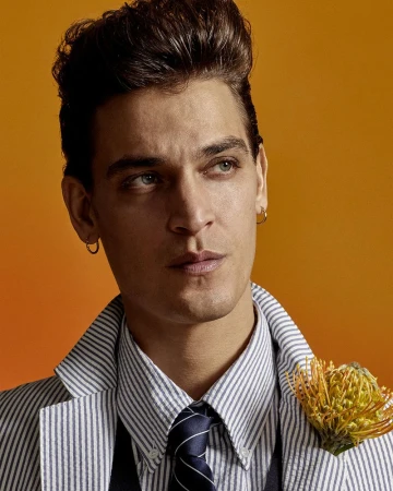 Adult male in a pinstripe jacket and navy tie poses in front of an orange gradient background with a flower in his lapel.