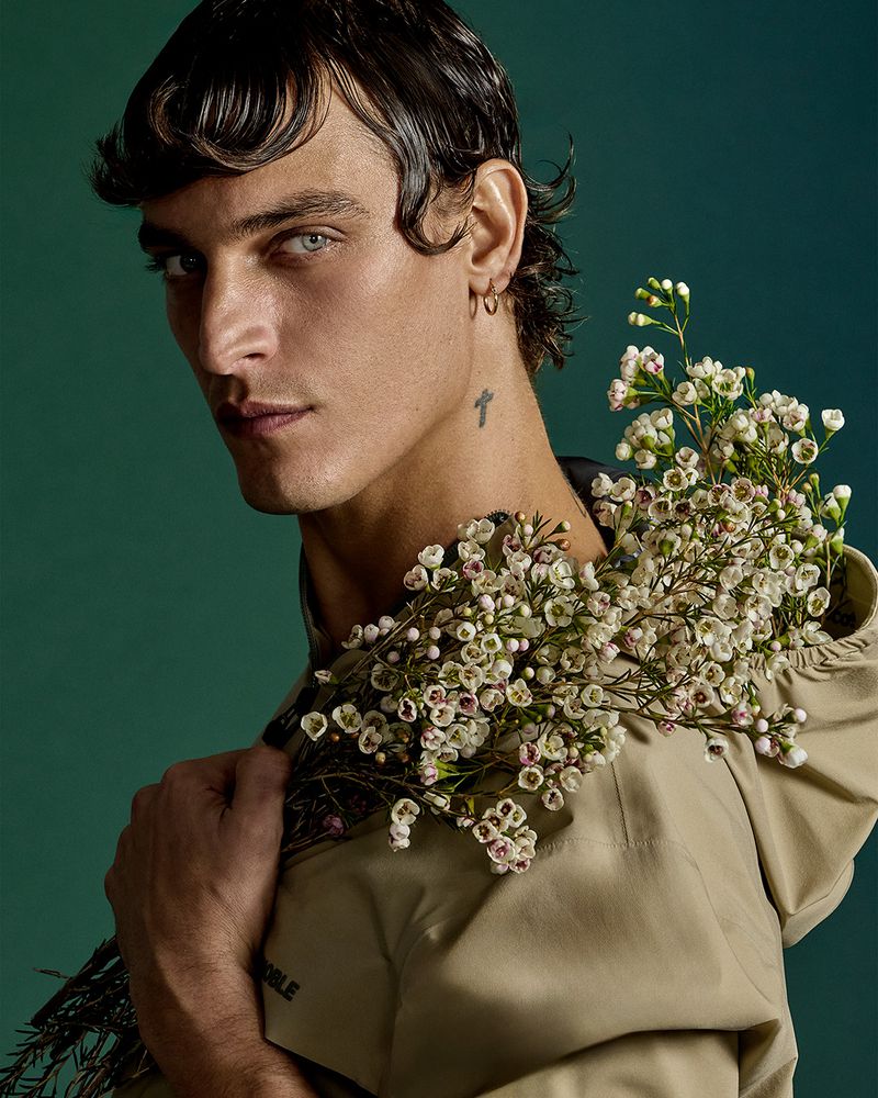 Model Jonathan Bellini holds a bunch of small white waxflowers over his shoulder against a solid green studio backdrop.