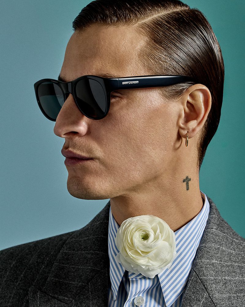 Portrait of an adult man wearing sunglasses, a striped shirt, and a grey jacket, accented with a white flower at the collar.