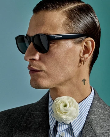 Portrait of an adult man wearing sunglasses, a striped shirt, and a grey jacket, accented with a white flower at the collar.  