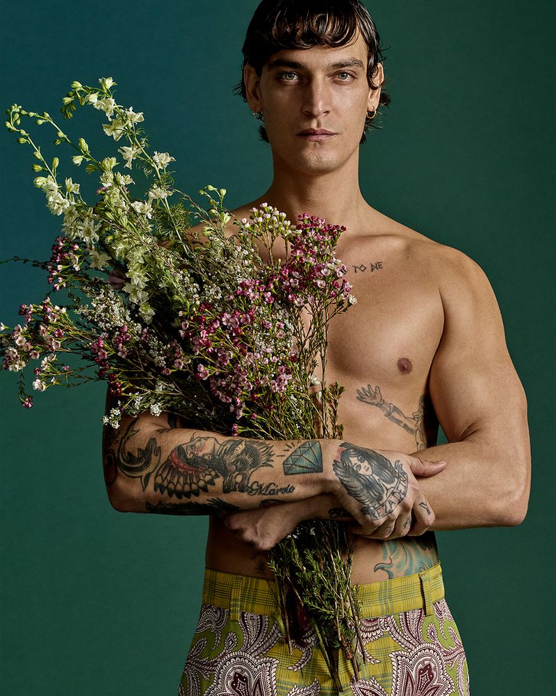 Shirtless male fashion model with tattoos holds a bouquet of colorful flowers against a teal studio backdrop in a portrait shot.