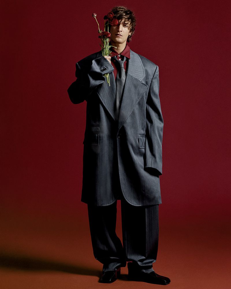 Young man in an oversized grey suit holds a bouquet of red flowers in a studio fashion portrait against a dark red background.