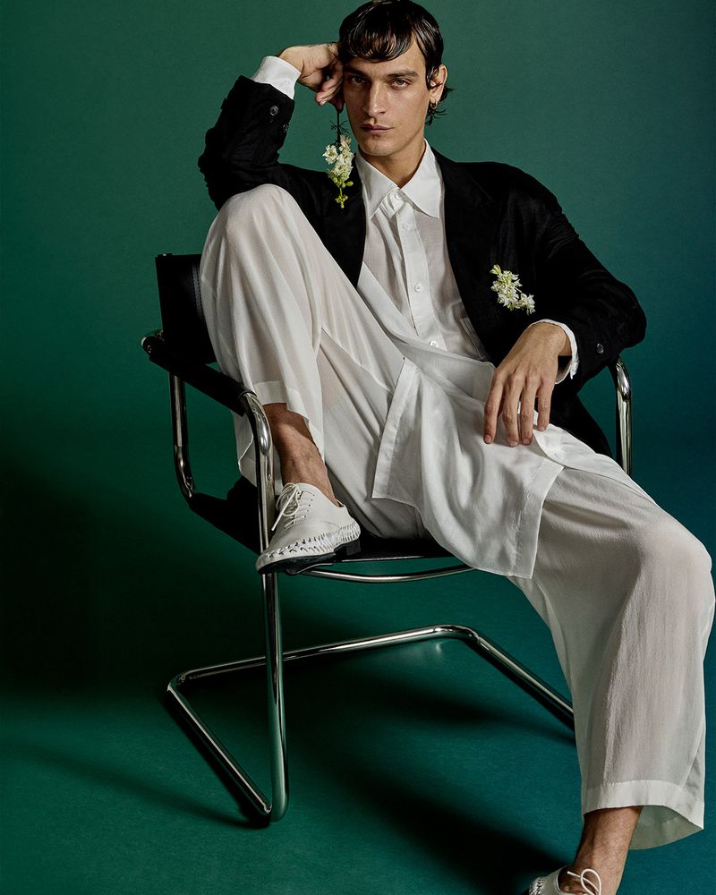 Male model in a black jacket and white pants seated in a tubular chair against a green backdrop in a fashion studio setting.