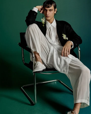 Male model in a black jacket and white pants seated in a tubular chair against a green backdrop in a fashion studio setting.