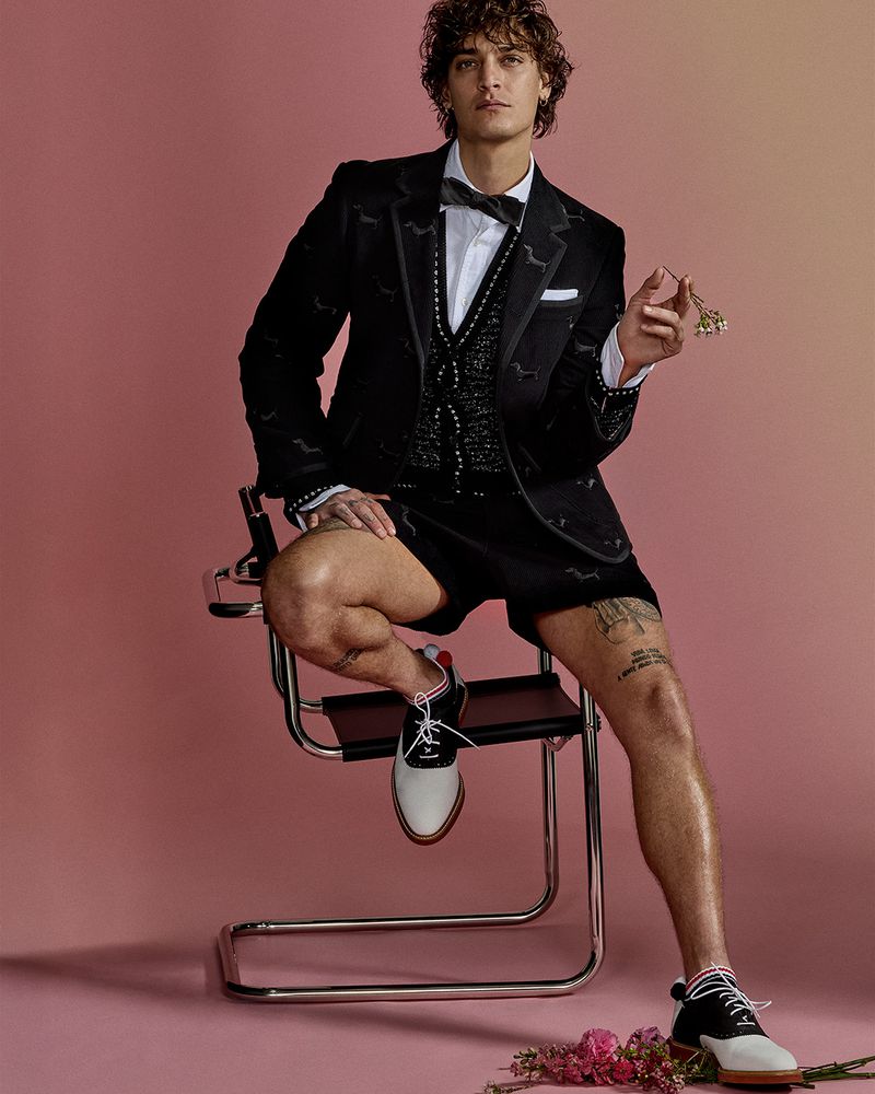 Male fashion model in a black tuxedo jacket, vest, and shorts sitting on a chair against a gradient pink background in a studio shot.