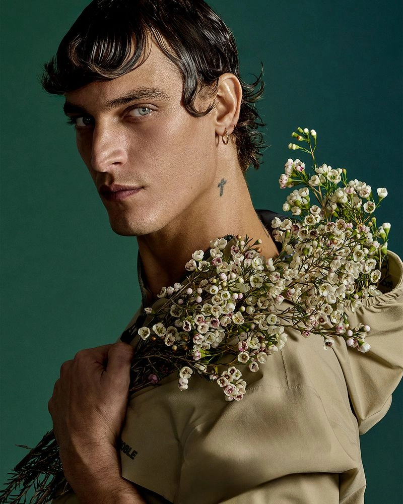 Model Jonathan Bellini holds a bunch of small white waxflowers over his shoulder against a solid green studio backdrop.