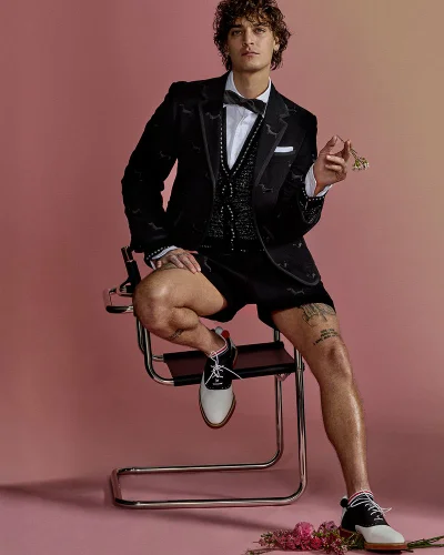 Model Jonathan Bellini seated in a black suit jacket shorts and saddle shoes against a warm gradient studio backdrop for Harper's Bazaar China.