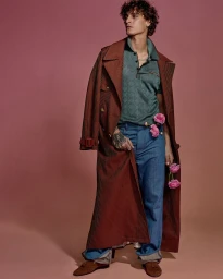 Full-length portrait of male model Jonathan Bellini wearing a striped trench coat and green polo shirt against a solid background