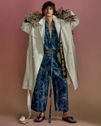 Model Jonathan Bellini wearing a white trench coat and blue patterned set with flowers behind shoulders against a warm gradient background.