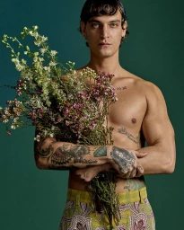 Model Jonathan Bellini shirtless with arm tattoos holding a bouquet of wildflowers against a teal green backdrop.