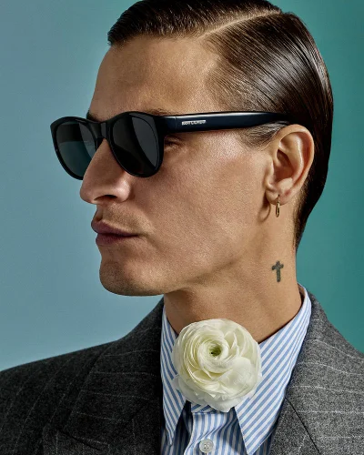 Close-up portrait of model Jonathan Bellini wearing black sunglasses, a pinstripe shirt with a white flower, and a grey blazer against a teal background.