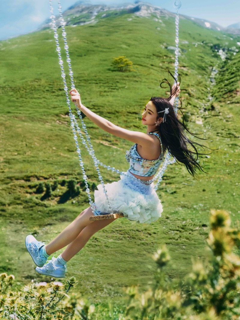 Woman swinging on a crystal-beaded swing above a green mountain slope wearing a ruffled skirt and blue sneakers.