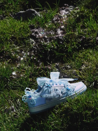 Light blue sneaker adorned with beaded flower embellishments and sheer ribbon laces lying on wet sunlit grass.