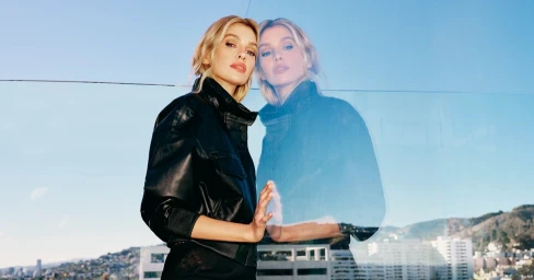Stella Maxwell in a black leather jacket reflected against a glass barrier with distant hills and buildings below
