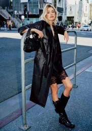 Stella Maxwell leaning against a metal street railing in a black leather coat and boots