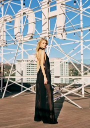 Stella Maxwell in a black backless gown on a rooftop with a large white metal structure against a blue sky.