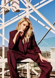 Stella Maxwell in a burgundy pantsuit posing against white structural framework under a clear blue sky
