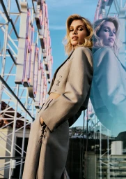Stella Maxwell in a taupe cropped blazer and matching skirt leaning against a reflective glass surface outdoors