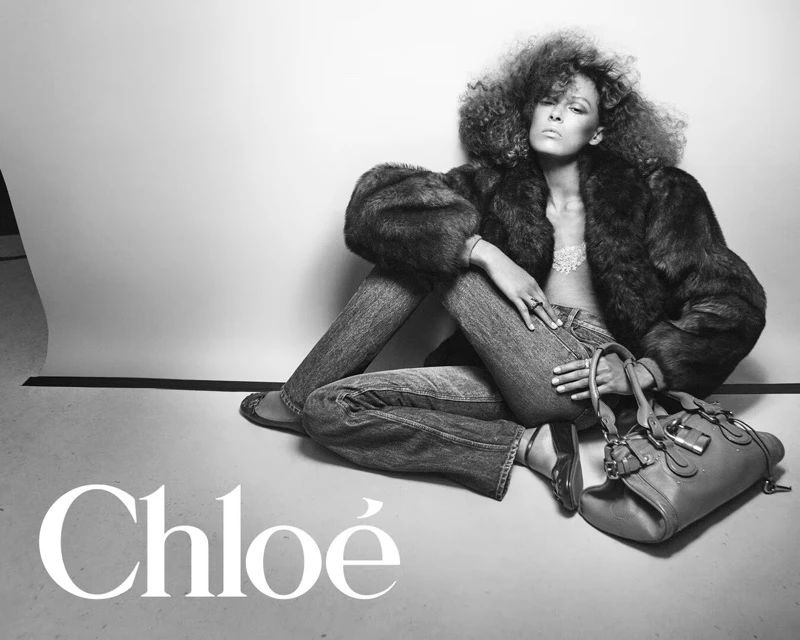 Model seated on a studio backdrop wearing a fur jacket and jeans next to a leather handbag, Chloé campaign.