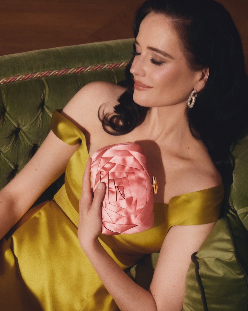 Eva Green in an off-the-shoulder yellow satin dress reclining on a green velvet sofa with a pink satin rose clutch bag.