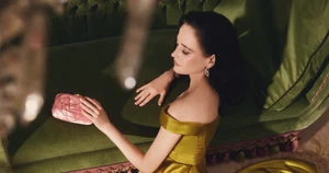 Eva Green wearing a yellow off-shoulder gown holding a pink clutch on a green velvet sofa for Roger Vivier
