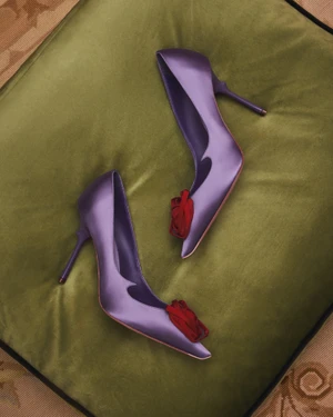 Pair of purple satin high heels with red rosette embellishments resting on a green velvet cushion.