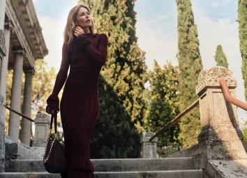 Doutzen Kroes in a burgundy knit dress carrying a dark shoulder bag on stone outdoor stairs with tall cypress trees in the background.
