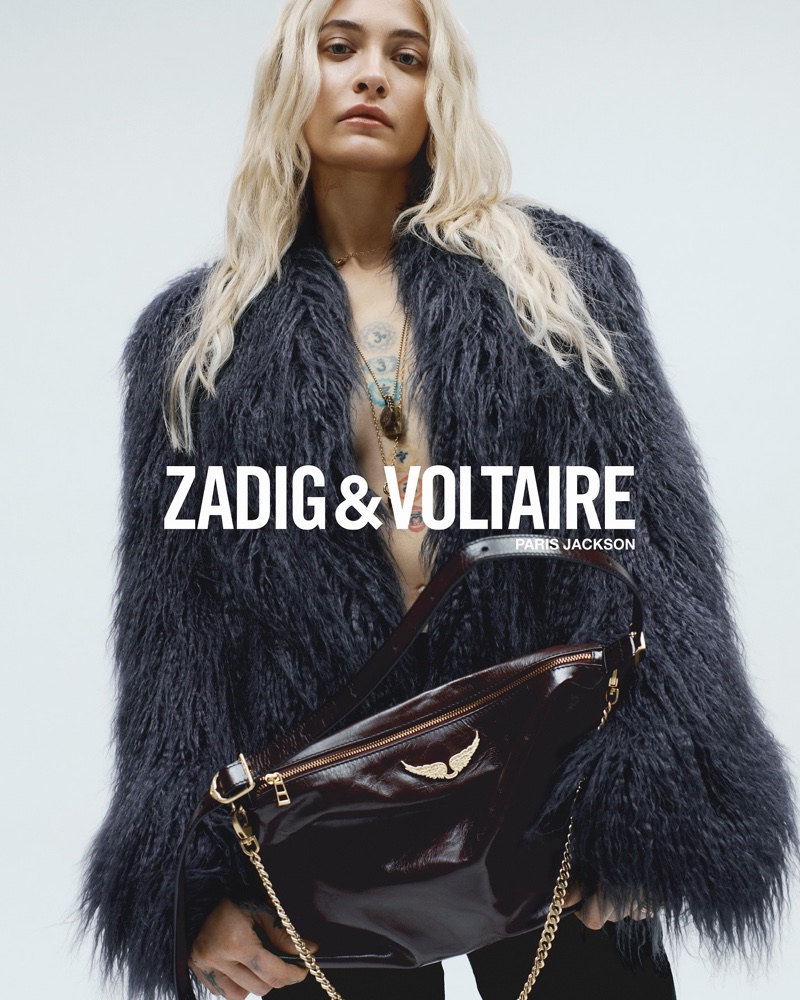 Paris Jackson wearing a dark shaggy fur coat and holding a burgundy leather shoulder bag against a white studio background.