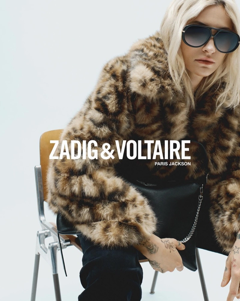 Paris Jackson wearing a faux fur leopard print coat and sunglasses, seated on a metal chair against a plain white studio background.