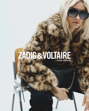 Paris Jackson wearing a faux fur leopard print coat and sunglasses, seated on a metal chair against a plain white studio background.