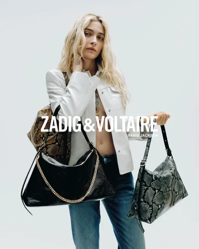 Paris Jackson holding multiple designer handbags wearing a white leather jacket and blue jeans against a white seamless backdrop