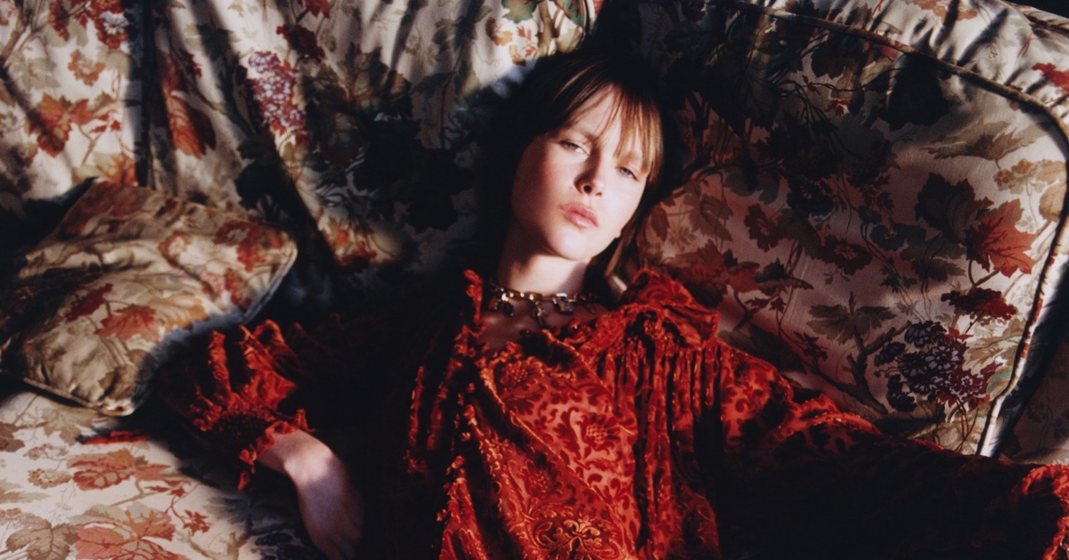 Model lying on a floral patterned sofa wearing a textured red patterned dress with ruffled neckline and gold chain necklace