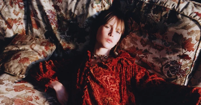 Model lying on a floral patterned sofa wearing a textured red patterned dress with ruffled neckline and gold chain necklace