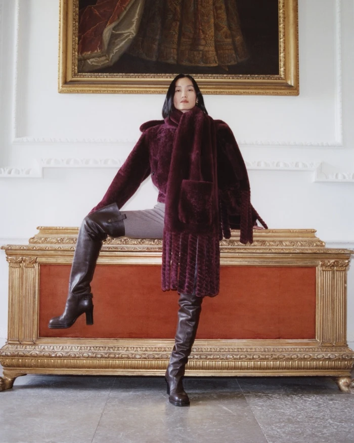 Model in burgundy faux fur coat and knee-high leather boots posing on a gilded vintage chest in an interior setting.