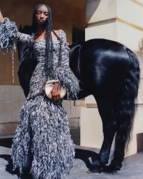 Model in a textured grey fringed dress posing next to a black horse outdoors against a classical stone building.