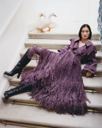Model in purple fringed trench coat and black boots reclining on marble stairs with white ducks behind her