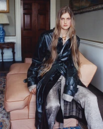 Model with long brown hair wearing a black leather coat seated on a peach sofa inside a room