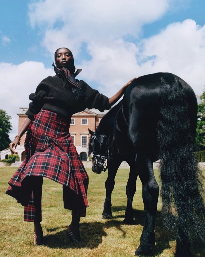 Model standing outdoors next to a black horse wearing a dark sweater and red plaid skirt with a stately building in the background.