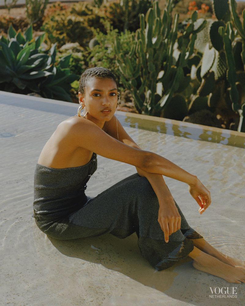 Woman with short hair sitting in water, wearing a grey jumpsuit and gold hoop earrings, with cacti in the blurred background.