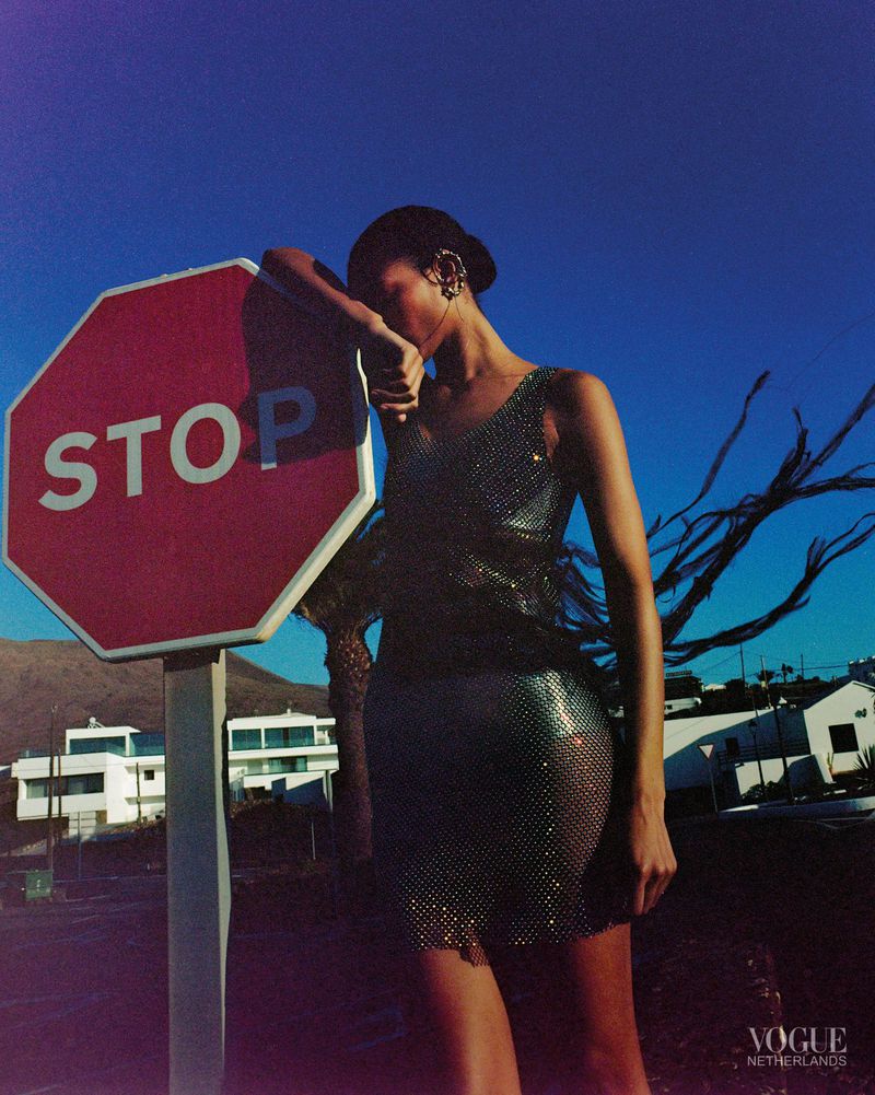 Fashion shot of a female model wearing a silver dress leaning against a red stop sign under a clear blue sky in an outdoor setting.