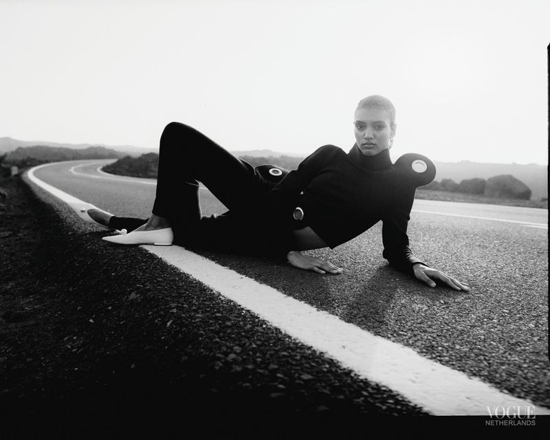 A model in a black outfit poses on a road under a clear sky with rolling hills in the background in a Vogue Netherlands fashion shot.