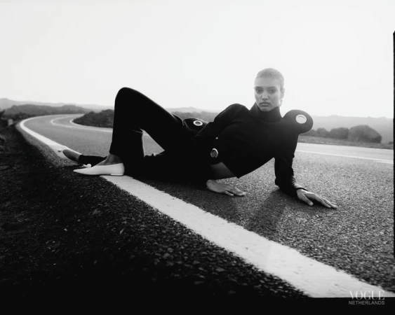 A model in a black outfit poses on a road under a clear sky with rolling hills in the background in a Vogue Netherlands fashion shot.