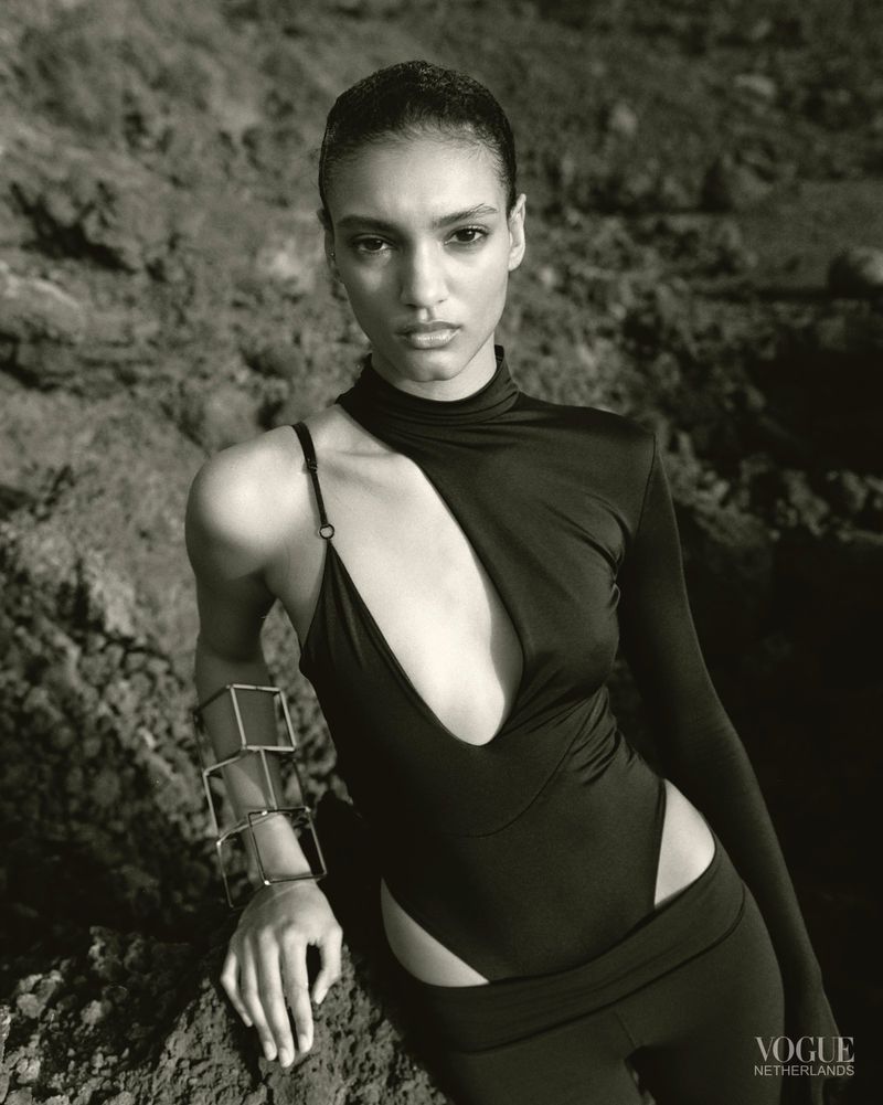 A model in a black bodysuit with a geometric cut-out poses against a rocky background, wearing a boxy metal cuff bracelet.