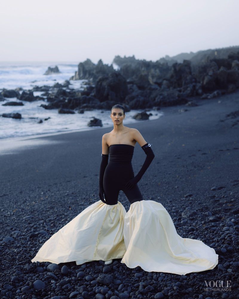 Fashion photography of a woman in a strapless black top and white skirt posing on a black sand beach with volcanic rocks.