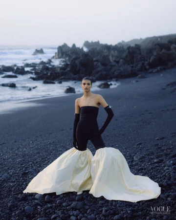 Fashion photography of a woman in a strapless black top and white skirt posing on a black sand beach with volcanic rocks.