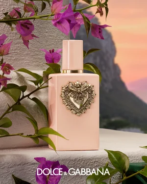 Pink Dolce and Gabbana perfume bottle with an ornate gold heart emblem resting outdoors among vibrant purple bougainvillea flowers