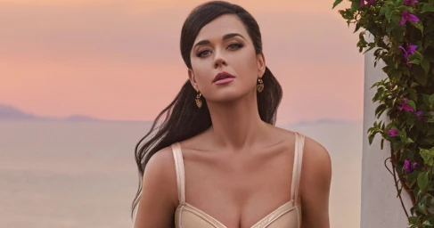 Katy Perry wearing a satin top with gold earrings against a coastal sunset background for Dolce and Gabbana campaign.