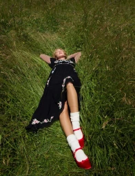 Model Dorit Revelis lying in tall green grass wearing a black floral dress, white socks, and red shoes